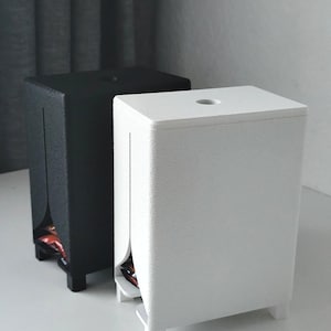 Puede incluir: Dos dispensadores rectangulares, uno negro mate y otro blanco mate, cada uno con una superficie texturizada, una abertura circular en la parte superior y una ranura vertical en el panel frontal que revela una peque&ntilde;a abertura en la base.