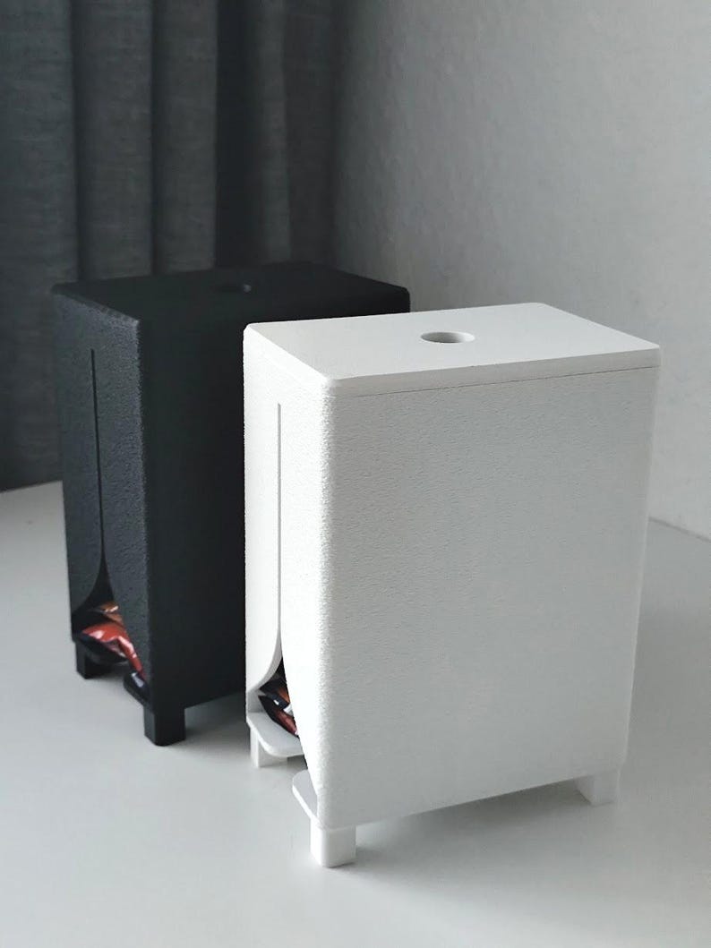 Puede incluir: Dos dispensadores rectangulares, uno negro mate y otro blanco mate, cada uno con una superficie texturizada, una abertura circular en la parte superior y una ranura vertical en el panel frontal que revela una peque&ntilde;a abertura en la base.