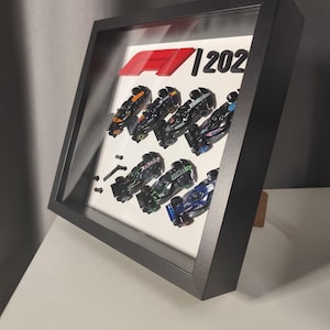 Peut inclure: Œuvre encadrée représentant des voitures de course de Formule 1 miniatures. Les voitures sont disposées sur un fond blanc, avec le logo F1 et l'année 2021. Le cadre est noir.
