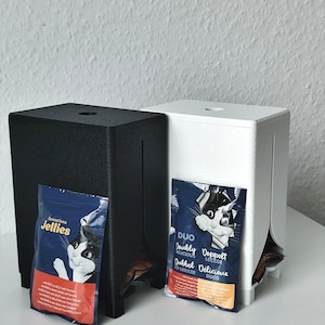 Puede incluir: Dos dispensadores rectangulares, uno negro y otro blanco, cada uno con una ranura vertical y una abertura superior. Una bolsa azul de comida para gatos con la etiqueta Sensations Jellies est&aacute; unida al dispensador negro, y una bolsa Duo al blanco.