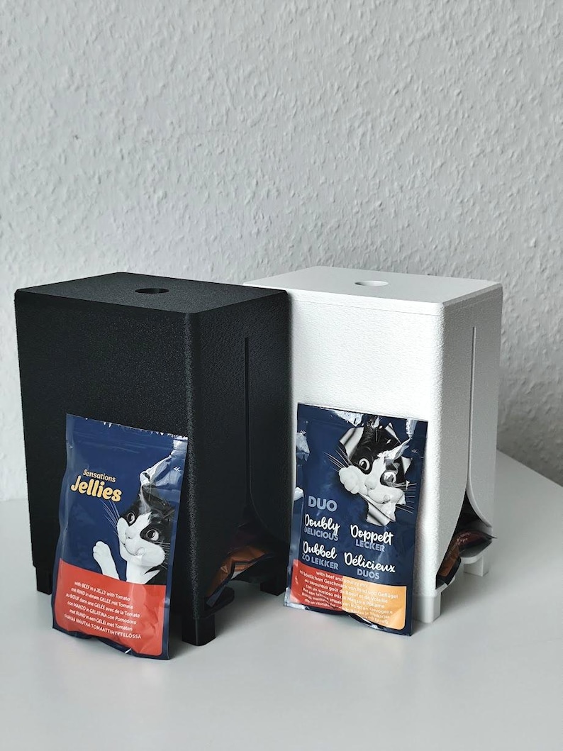 Puede incluir: Dos dispensadores rectangulares, uno negro y otro blanco, cada uno con una ranura vertical y una abertura superior. Una bolsa azul de comida para gatos con la etiqueta Sensations Jellies est&aacute; unida al dispensador negro, y una bolsa Duo al blanco.