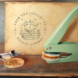 Könnte beinhalten: Ein grüner Stempel mit Selbsttinte und einem goldenen Siegel, das eine Fledermaus und ein Buch mit dem Text "From the Library of Breeze Alvarez" zeigt.