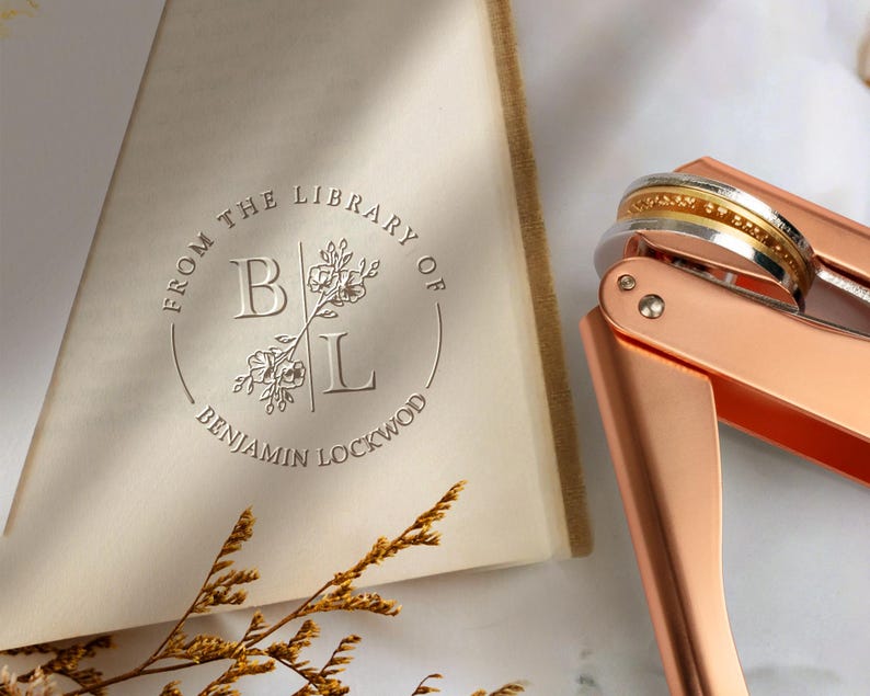 Peut inclure: Un gaufrage de livre en or rose avec une bague en or et argent, &agrave; c&ocirc;t&eacute; d'une page de livre. La page est estamp&eacute;e du texte "FROM THE LIBRARY OF BENJAMIN LOCKWOOD" autour des lettres "BL" et d'&eacute;l&eacute;ments floraux. Le livre a une couverture beige clair.