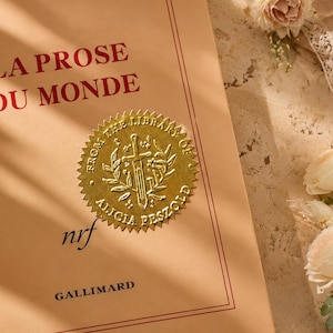 Peut inclure: Un livre intitul&eacute; "LA PROSE DU MONDE" avec du texte rouge sur une couverture beige clair. Un sceau dor&eacute; avec une &eacute;p&eacute;e et une couronne est appos&eacute; sur la couverture. Le livre est de la biblioth&egrave;que d'Alicia Peszold, publi&eacute; par Gallimard.