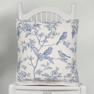 May include: A square decorative pillow with a blue and white toile design featuring birds perched on branches. The pillow is placed on a white chair. The pillow has a floral and bird pattern.