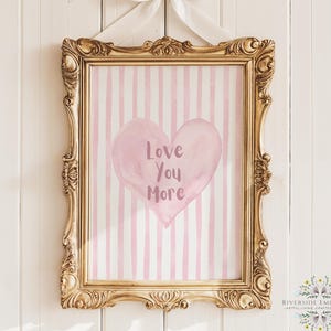 May include: A framed print with a gold ornate frame and a white bow. The print features a pink heart with the words "Love You More" on a pink and white striped background. The frame is hanging on a white wall.