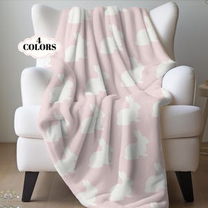 May include: A soft, light pink fleece throw blanket featuring a repeating pattern of white rabbit silhouettes. The blanket is draped over a white armchair. A small white graphic badge on the upper left side displays the text 4 COLORS.