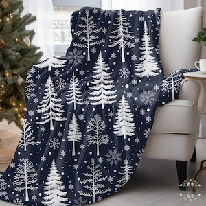 Könnte beinhalten: Eine dunkelblaue Wohndecke mit einem sich wiederholenden Muster aus weißen Tannenbäumen und Schneeflocken. Die Decke liegt über einem weißen Sessel und zeigt eine weiche Textur mit einem durchgehenden winterlichen Druck auf der gesamten Stoffoberfläche.