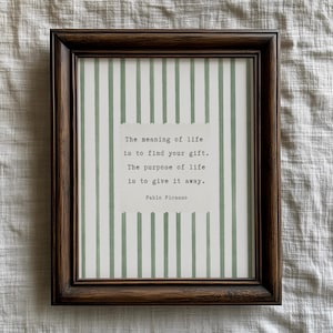 May include: A framed print with a dark brown wooden frame. The print features a white background with vertical green stripes. The text reads: "The meaning of life is to find your gift. The purpose of life is to give it away." by Pablo Picasso.