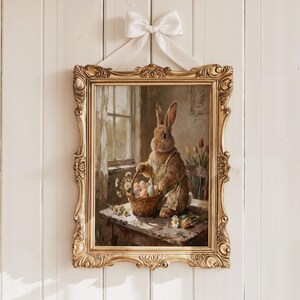 May include: A rectangular wall art piece in an ornate gold frame with a white ribbon bow at the top. The print depicts a brown rabbit wearing an apron, standing at a wooden table with a basket of colorful eggs and white daisies near a window.