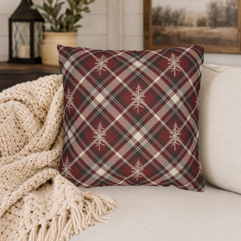 May include: A square throw pillow featuring a diagonal plaid pattern in shades of deep red, dark green, and cream. The fabric is decorated with a repeating print of small, light-colored pine tree silhouettes centered within the plaid intersections.