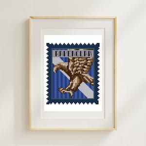 May include: A framed Ravenclaw cross-stitch art piece. The artwork features a brown eagle emblem on a blue and white striped background, resembling a postage stamp. The word "RAVENCLAW" is displayed at the top. The frame is a light wood color.