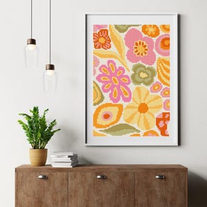 May include: A framed floral print featuring a retro design with pink, orange, yellow, and green flowers. The artwork is displayed on a white background and framed in a black frame. The print is displayed above a wooden cabinet.