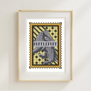 May include: Framed cross-stitch art featuring a yellow and black stamp design with the text "HUFFLEPUFF" in white. The stamp has a gray image and is set against a yellow and black patterned background. The frame is a light wood color.