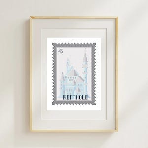 May include: A framed print featuring a cross-stitch design of a light blue and white castle. The design is in the shape of a postage stamp with the word "RIETHOLD" and the number "45" visible. The frame is a light wood color.