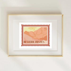 May include: Framed cross-stitch art featuring a postage stamp design with a landscape scene in shades of peach, orange, and yellow. The stamp reads "WESTERN WASTES" and has a value of "60". The artwork is displayed in a light wood frame.