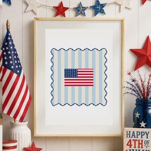 May include: A framed cross-stitch pattern featuring a central United States flag design on a light blue and white vertical striped background with a scalloped dark blue border, presented in a simple light-colored wooden frame.