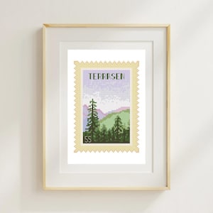 May include: A framed cross-stitch artwork featuring a landscape design resembling a postage stamp. The design includes green trees, purple mountains, and the word "TERRASEN". The artwork is framed in a light wood frame.