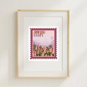 May include: A framed print featuring a postage stamp design with the words "SPRING COURT" and the number "14". The stamp depicts a pixelated landscape in shades of pink, green, and white, framed in a dark pink border. The frame is a light wood color.
