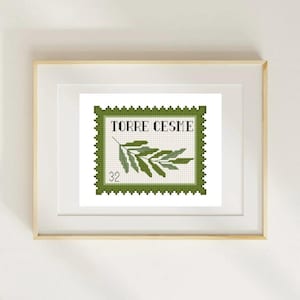 May include: A framed cross-stitch artwork featuring a green and white design resembling a postage stamp. The stamp reads "TORRE CESME" and depicts a sprig of leaves. The number "32" is also visible. The frame is a light wood color.
