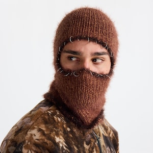 May include: A brown knitted balaclava with silver rings around the face opening. The balaclava is made of a fuzzy, textured yarn. The person in the image is wearing a brown and tan patterned jacket.