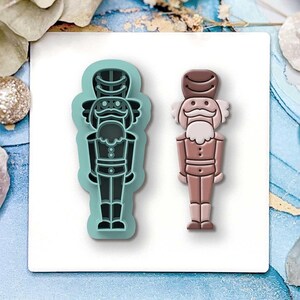 May include: Two nutcracker-shaped cookie cutters. One is a teal outline, and the other is a light brown colour. The nutcrackers have a hat, moustache, and uniform. The cutters are on a white surface with holiday decorations in the background.