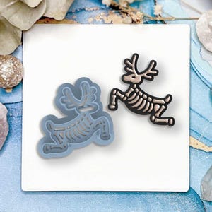 以下が含まれることがあります： 2つのトナカイの骨格デザイン。1つは水色の型で、もう1つは黒とベージュの完成品です。トナカイは走っているポーズで、角と肋骨が見えます。画像は白い正方形にあります。