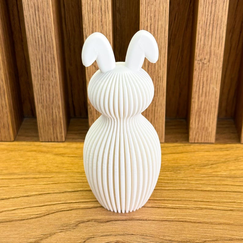 May include: White ceramic bunny figurine with a ribbed body and long ears. The figurine is standing on a wooden surface.