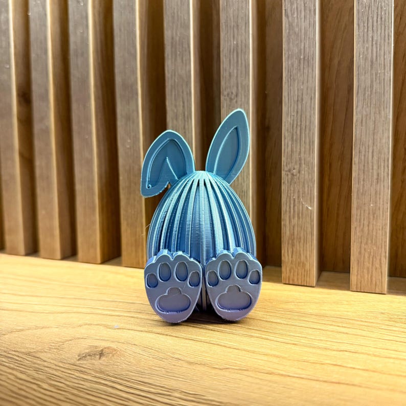 May include: A blue 3D printed Easter bunny egg with bunny ears and feet. The egg is decorated with a ribbed pattern.