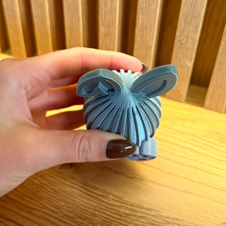 May include: A light blue 3D printed Easter egg with bunny ears. The egg has a textured surface and is decorated with a pattern of lines.