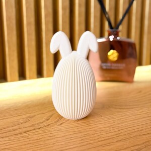 May include: White ceramic bunny-shaped egg with a ribbed texture. The egg has two small bunny ears on top.