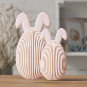 May include: Two pink ceramic bunny-shaped Easter eggs. The larger egg is in the front and the smaller egg is behind it. Both eggs have a ribbed texture.