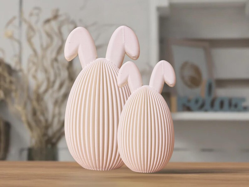 May include: Two pink ceramic bunny-shaped Easter eggs. The larger egg is in the front and the smaller egg is behind it. Both eggs have a ribbed texture.