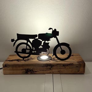 Op de afbeelding: Een zwarte motorfiets-silhouetlamp op een rechthoekige houten basis. De lamp heeft een kleine, ronde lichtbron. De houten basis heeft een natuurlijke, rustieke uitstraling. De lamp is aangesloten op een stopcontact.