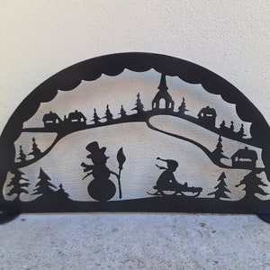 Könnte beinhalten: Schwarze Metallsilhouette einer Winterszene mit einem Schneemann, einer Person, die Schlitten fährt, und einer Kirche im Hintergrund. Die Silhouette hat die Form eines Halbkreises und hat zwei Metallfüße.