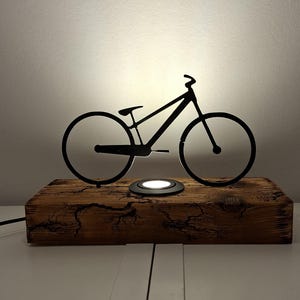 Puede incluir: Una lámpara con silueta de bicicleta de metal negro se asienta sobre una base de madera rústica y desgastada. La lámpara presenta una luz circular y un diseño elegante y moderno. El fondo es una pared neutra.