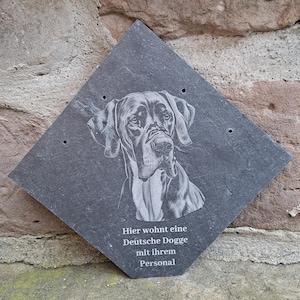 May include: A dark grey, diamond-shaped slate sign with an engraved portrait of a Great Dane. The sign includes the German text "Hier wohnt eine Deutsche Dogge mit ihrem Personal."