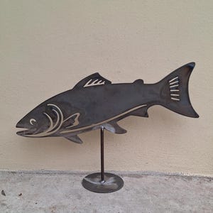 May include: A metal sculpture of a fish, standing on a round metal base. The fish is a dark gray color and has a detailed design.