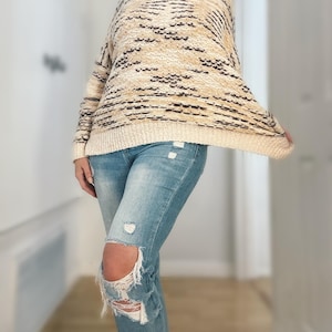 May include: A cream-colored sweater with a textured pattern in shades of tan and black. The sweater is paired with light blue distressed jeans. The jeans have ripped knees.
