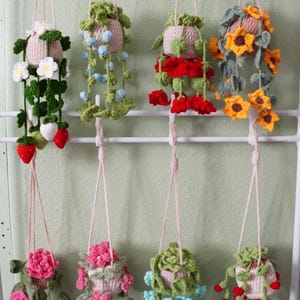May include: A collection of handmade crochet hanging planters, each featuring a unique floral design. The planters are suspended by pink cords and showcase various flowers, including white daisies, red strawberries, and yellow sunflowers. The background is a light green wall.