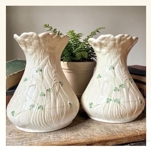 Puede incluir: Dos jarrones de cerámica blanquecina con un patrón vertical texturizado y detalles de tréboles verdes. Los jarrones tienen una parte superior acampanada y con volantes y una base bulbosa. Una pequeña planta en maceta es visible en el fondo.