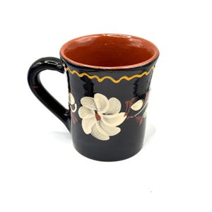 Könnte beinhalten: Eine schwarze Keramiktasse mit einem weißen Blumendesign und einem gelben Rand. Die Tasse hat ein braunes Inneres.