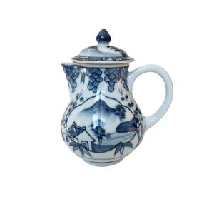 May include: A blue and white ceramic pitcher with a lid. The pitcher features a landscape scene with a mountain, trees, and a house. Grapes and vines decorate the body of the pitcher. The handle is white.