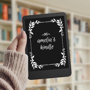 May include: A black e-reader with a white floral border and the text "amelia's kindle" in a cursive font. The e-reader is held in a hand, with a bookshelf in the background. The words "DEWDROPS OF COMFORT" are at the bottom.