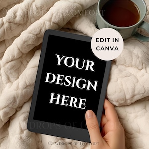 May include: A black tablet displays the text "YOUR DESIGN HERE" in white, held by a hand. The tablet rests on a cream-colored, textured blanket. A mug of tea sits nearby, and a circular "EDIT IN CANVA" sticker is visible.