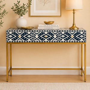 May include: A gold-legged console table with three drawers. The drawers are decorated with a navy and white geometric pattern. A gold lamp, artwork, and decorative items sit on top of the table. A vase with greenery and a woven basket are also visible.