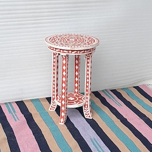 Può includere: Un tavolino rotondo rosso con dettagli in intarsio di osso bianco. Il tavolo ha un piano circolare e tre gambe.