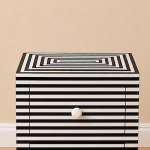 May include: A black and white striped nightstand with a geometric design on the top. The drawer features a white knob. The nightstand is a modern piece of furniture with a bold, graphic pattern.