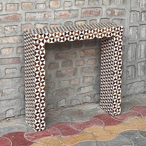 May include: A rectangular console table featuring a repeating geometric pattern of alternating dark brown and white triangles. The structure has a flat top surface and two vertical side panels, creating an open archway design.