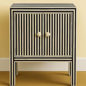 May include: A black and white striped cabinet with two doors and two round, off-white knobs. The cabinet has a rectangular shape and is supported by four legs. The design features vertical stripes and a contrasting border.